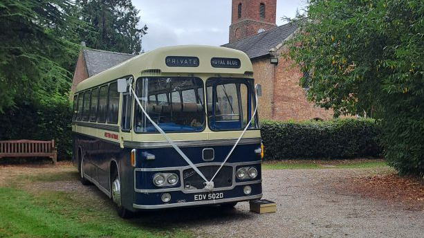 Front-on view of a blue and ivory vintage single-decker bus parked on a gravel drive surrounded by green bushes and trees.