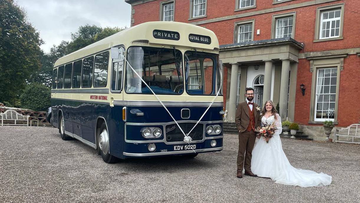 Blue and ivory vintage single-decker bus shown from a front angled view with a bride and groom standing beside it in front of a red-brick wedding venue.