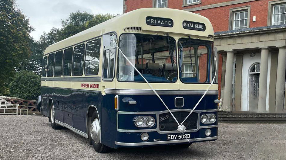 Blue and ivory vintage single-decker bus photographed from a slight front angle, parked outside a grand red-brick building with columns.