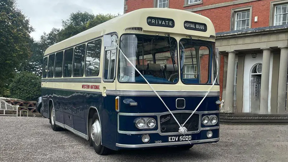 Blue and ivory vintage single-decker bus photographed from a slight front angle, parked outside a grand red-brick building with columns.