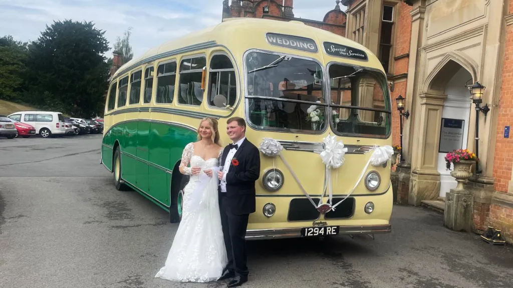 Yellow and green vintage single-decker wedding bus decorated with white bows, with a bride and groom standing beside it outside a manor house.