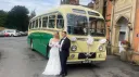 Yellow and green vintage single-decker wedding bus decorated with white bows, with a bride and groom standing beside it outside a manor house.