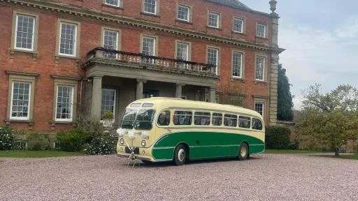 Yellow and green vintage single-decker bus photographed from a side angle outside a red-brick manor building.