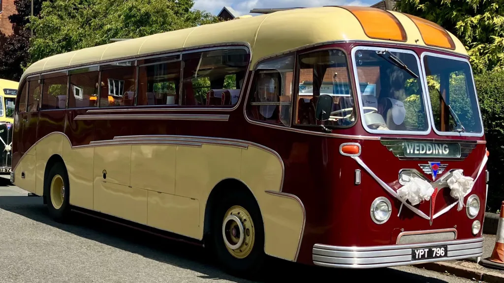 Side view of a burgundy and cream vintage single-decker bus parked on a sunny roadside with trees in the background.