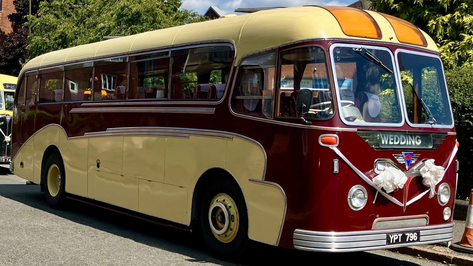 Side view of a burgundy and cream vintage single-decker bus parked on a sunny roadside with trees in the background.