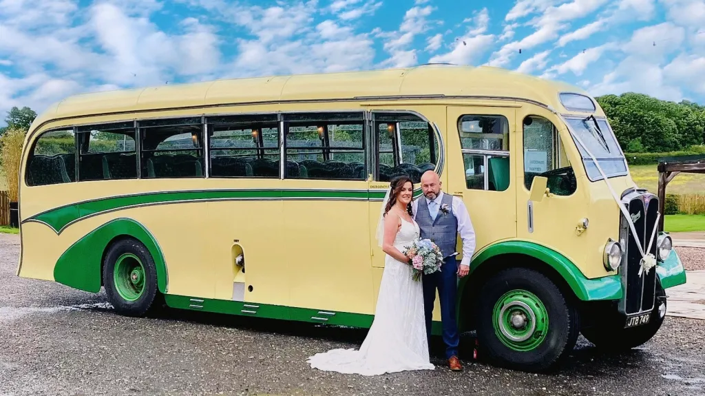 Yellow and green vintage single-decker bus captured from a side angle on a sunny day with the bride and groom posing next to it on a tarmac area.