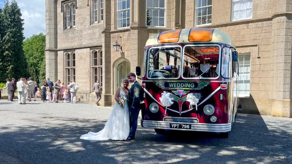 Front angled view of a burgundy and cream vintage bus parked outside a stone venue, with the bride and groom posing alongside it.