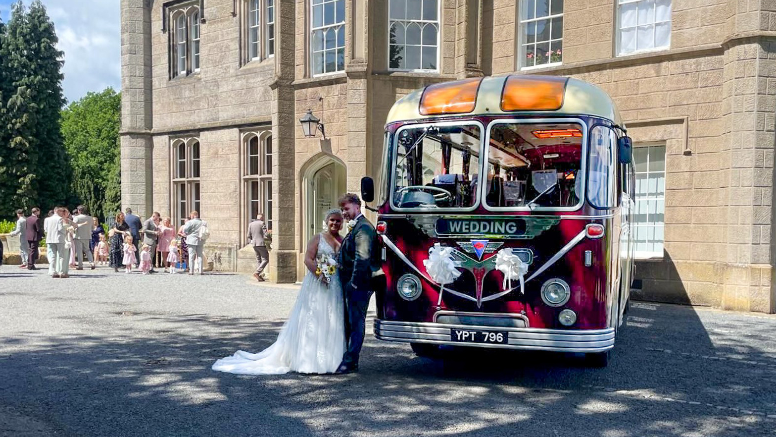 Front angled view of a burgundy and cream vintage bus parked outside a stone venue, with the bride and groom posing alongside it.