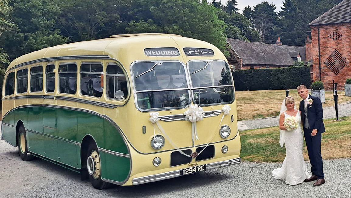 ellow and green vintage single-decker bus shown from the side with a bride and groom posing beside it on a sunny wedding day.