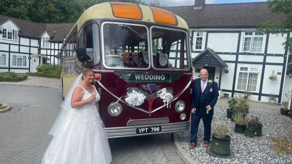 Vintage burgundy and cream wedding bus decorated with white bows, shown from a front angled view with a bride standing beside it in a courtyard.