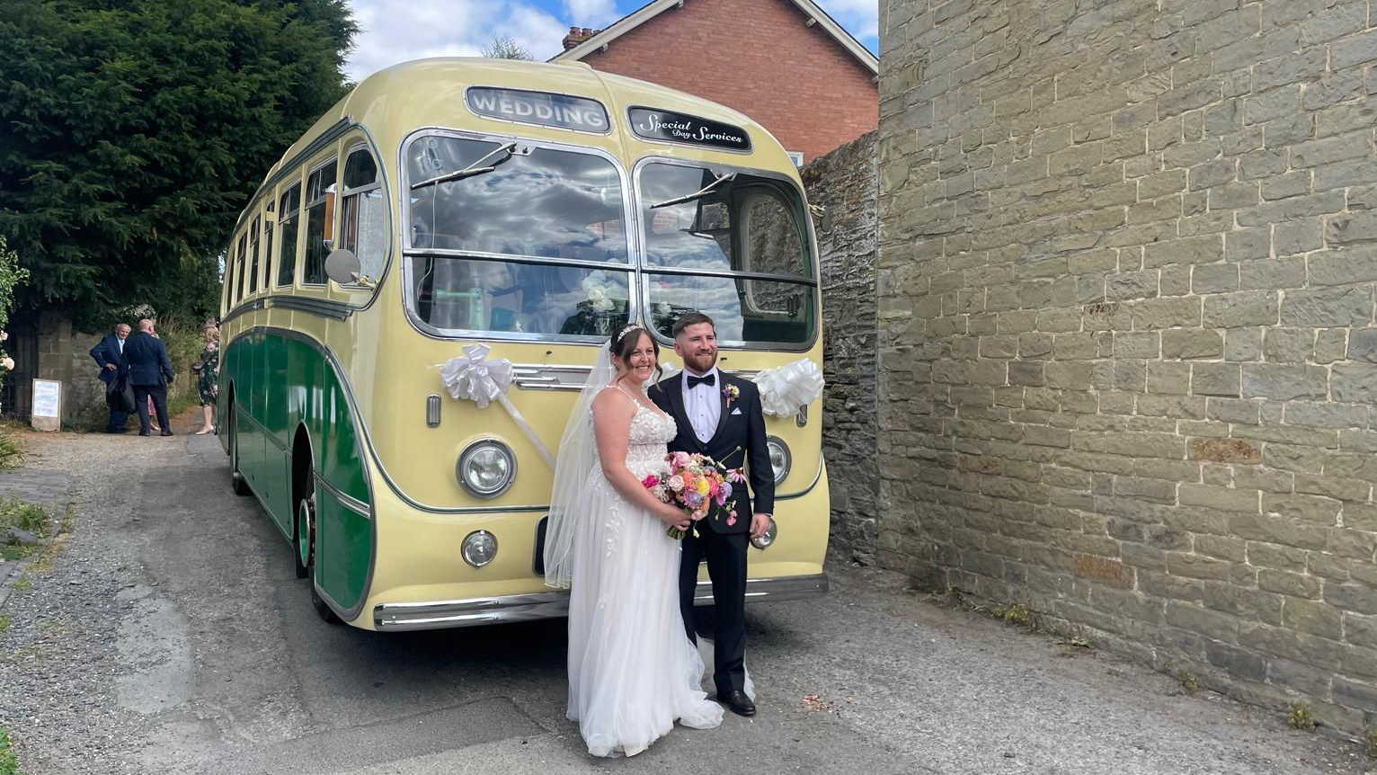 Yellow and green vintage single-decker bus photographed from the front angle with a bride and groom standing in front of a stone church wall.