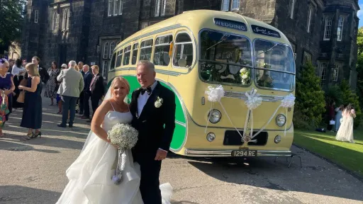 Yellow and green vintage single-decker bus shown from a side-front angle with a bride and groom posing in a churchyard area.
