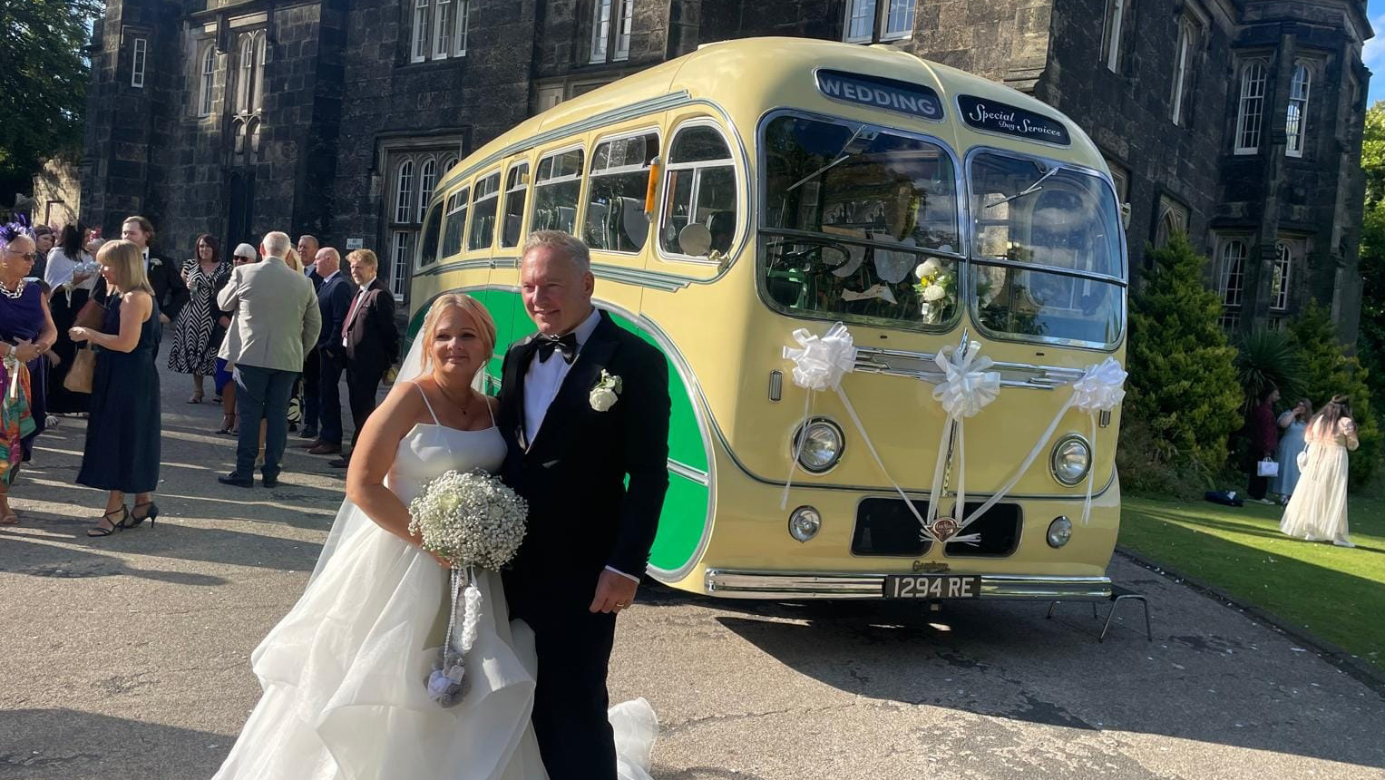 Yellow and green vintage single-decker bus shown from a side-front angle with a bride and groom posing in a churchyard area.