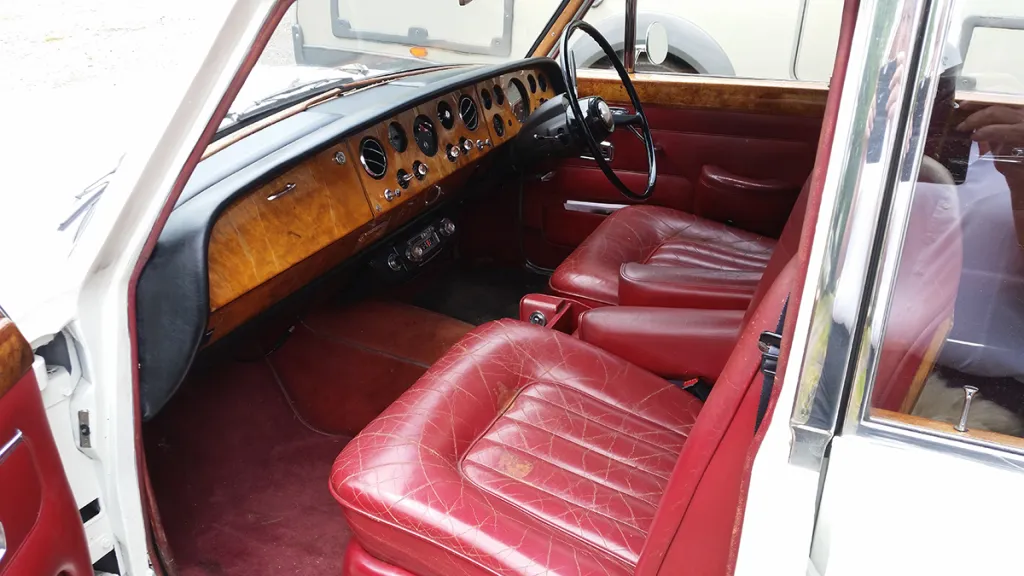 Front interior of a 1967 Rolls Royce Silver Shadow Mk I featuring deep red leather seats, polished wooden dashboard and classic chrome detailing.