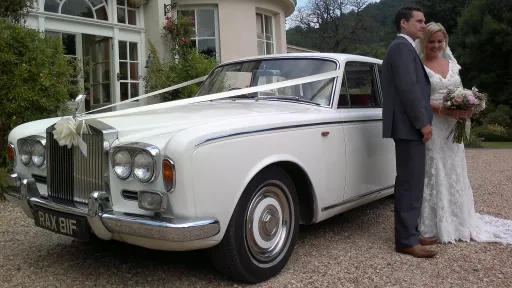 Bride and groom standing beside a Rolls Royce Silver Shadow Mk I in white, parked outside a wedding venue, showcasing its elegant 1960s body shape and chrome grille.
