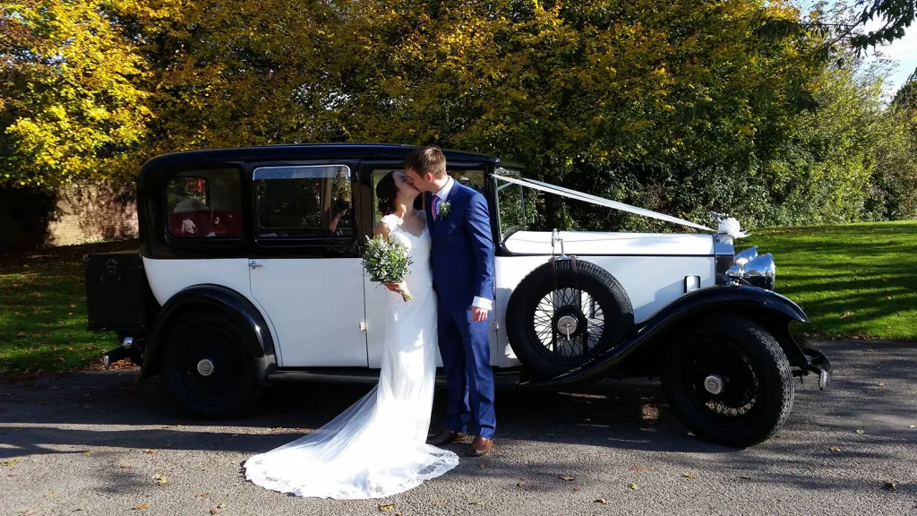 Bride and groom standing beside a vintage black and white Rolls Royce wedding car, surrounded by autumn trees and soft golden light.