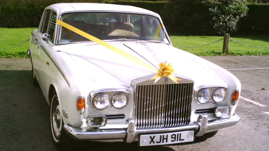 Front view of a white Rolls Royce Silver Shadow decorated with yellow wedding ribbons, showing the signature Rolls Royce grille and twin headlamps.