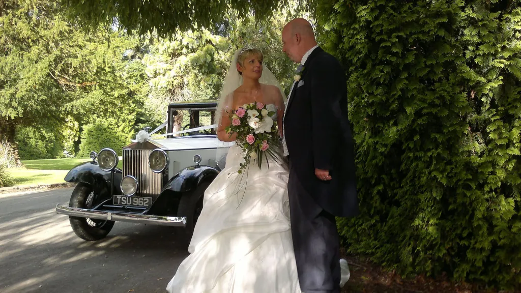 Smiling wedding couple posing in front of a vintage Rolls Royce in a leafy garden setting, with the car positioned behind them for photographs.