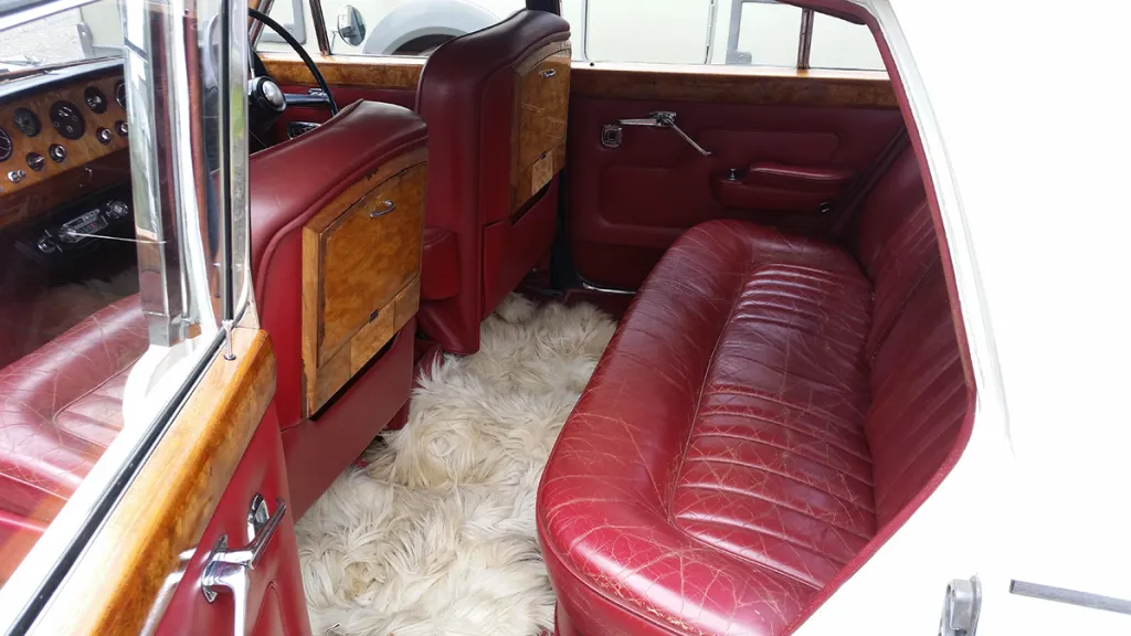 Rear interior view of a classic Rolls Royce Silver Shadow Mk I featuring plush red leather seating, soft floor rugs and polished wooden trim.