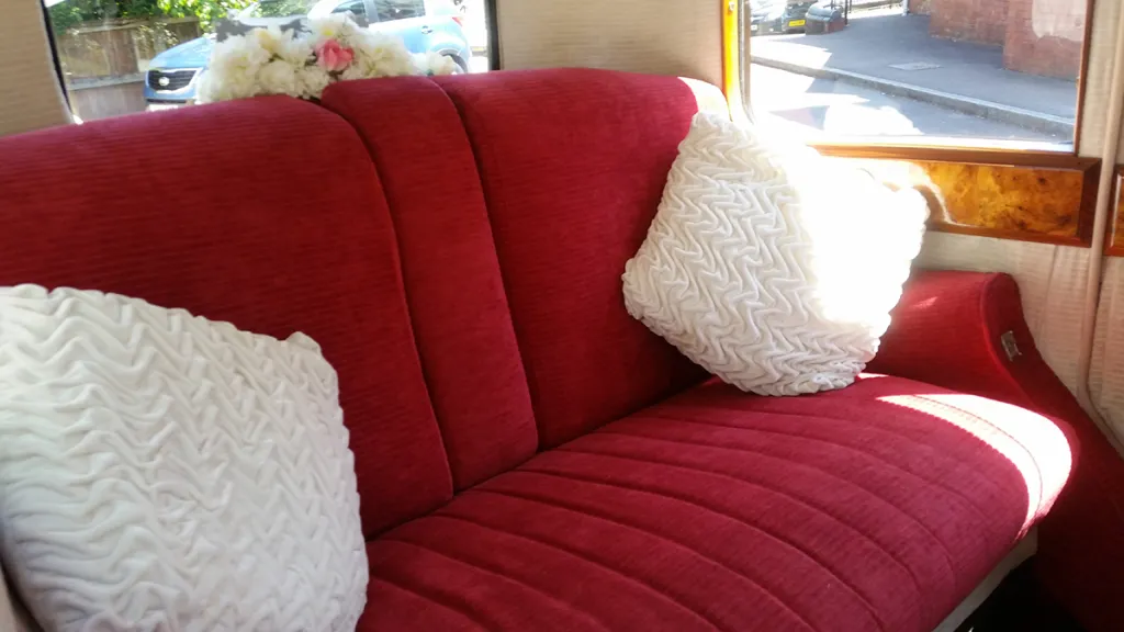 Close-up of the vintage Rolls Royce interior showing plush red velour seating with white cushions and classic wooden window surrounds.