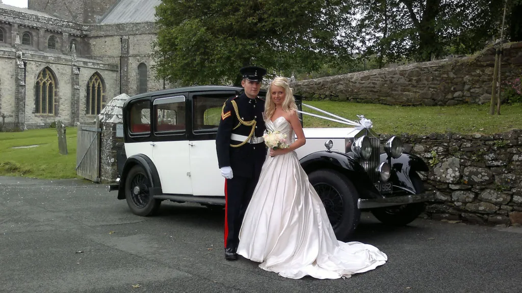 Vintage black and white Rolls Royce wedding car parked outside a church as the bride and groom pose beside it for their wedding photos.