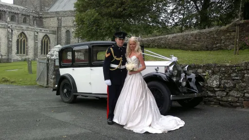 Vintage black and white Rolls Royce wedding car parked outside a church as the bride and groom pose beside it for their wedding photos.