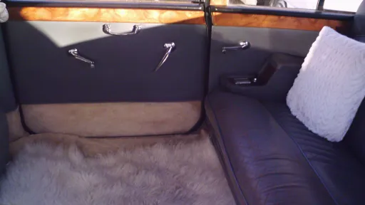 Interior of an 80s classic Daimler limousine showing tan leather seating, wooden trim and a white cushion on the passenger rear bench seat.