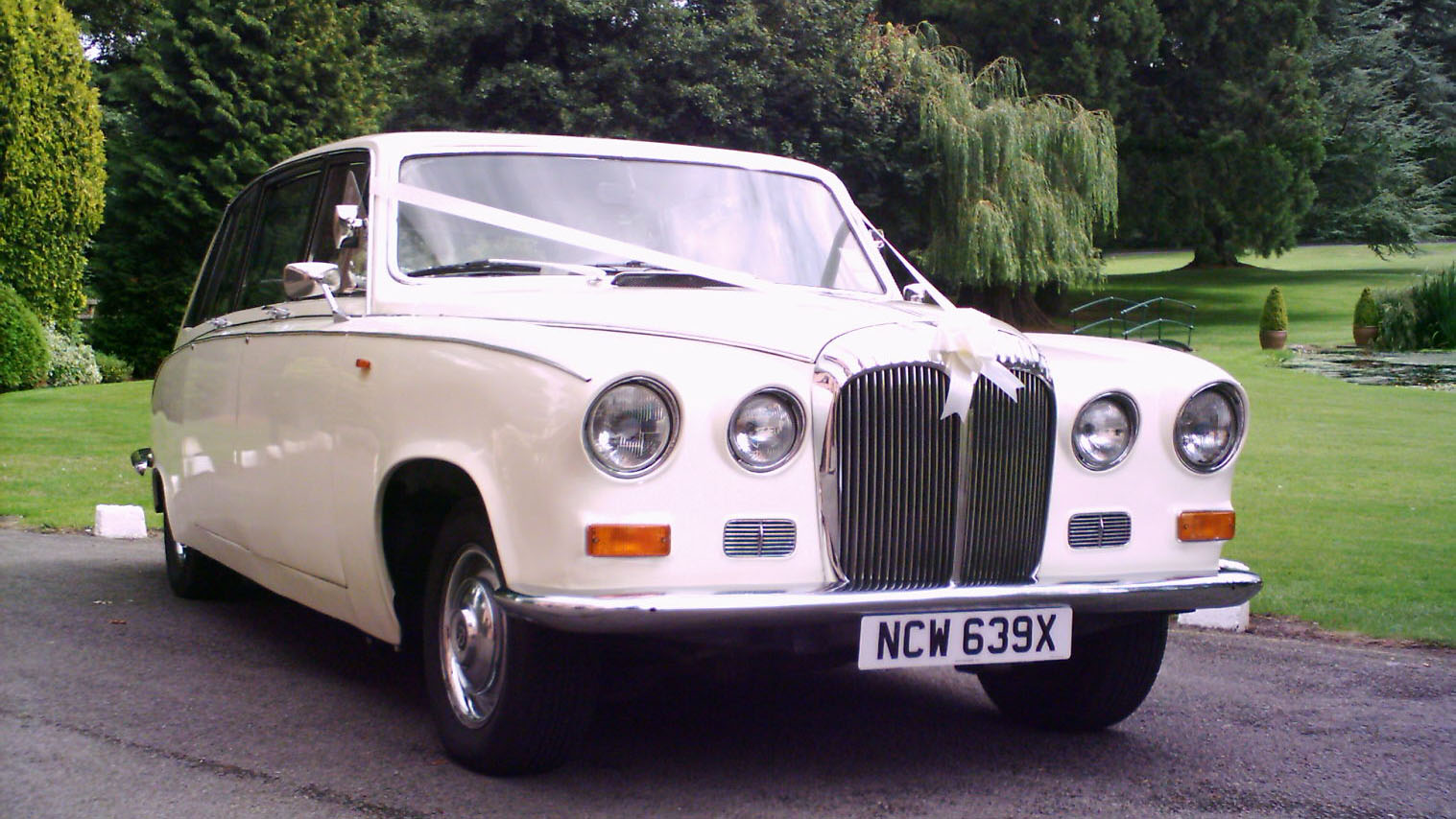 ront three-quarter view of a cream 1980s Daimler limousine parked on a driveway with greenery in the background.