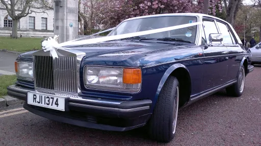 Front view of a 1990 blue Rolls-Royce Silver Spirit with a white vinyl roof, decorated with white wedding ribbons.