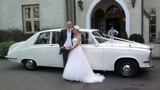Bride and groom standing beside a cream classic Daimler limousine decorated with white ribbons outside a grand entrance.