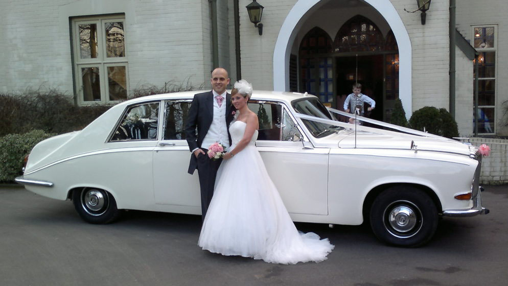 Bride and groom standing beside a cream classic Daimler limousine decorated with white ribbons outside a grand entrance.