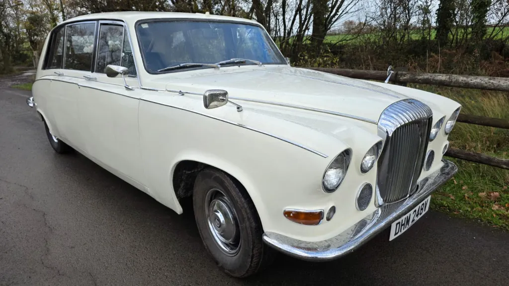 Front-side view of a cream classic Daimler limousine decorated with white wedding ribbons, parked on a country road.