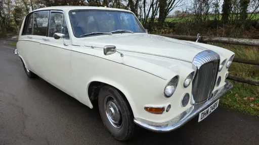 Front-side view of a cream classic Daimler limousine decorated with white wedding ribbons, parked on a country road.