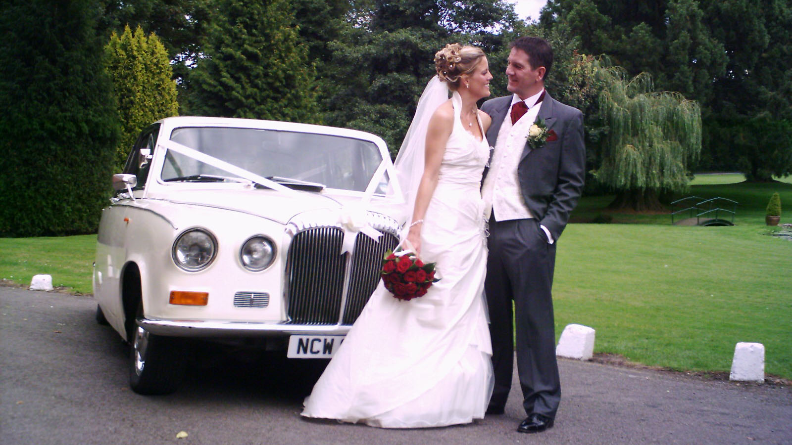 Cream classic Daimler limousine with wedding ribbons, parked on lawn with bride and groom standing beside the car.