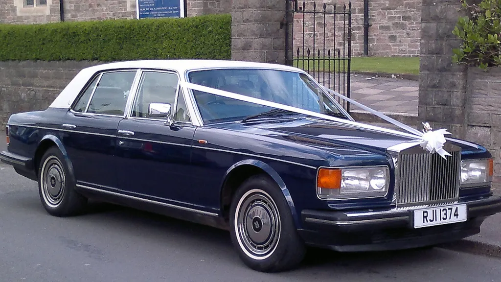 Side-front view of a blue Rolls-Royce Silver Spirit with a white roof and white wedding ribbons, parked beside a stone wall.