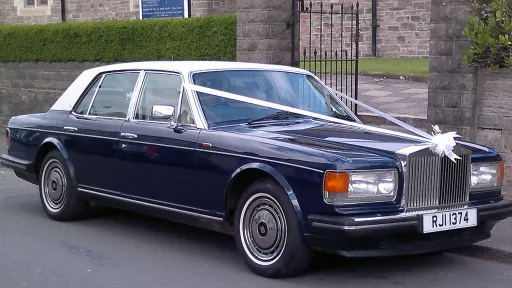 Side-front view of a blue Rolls-Royce Silver Spirit with a white roof and white wedding ribbons, parked beside a stone wall.