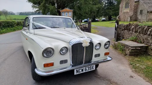 Front view of a cream classic Daimler limousine decorated with white wedding ribbons, parked outside a stone church.