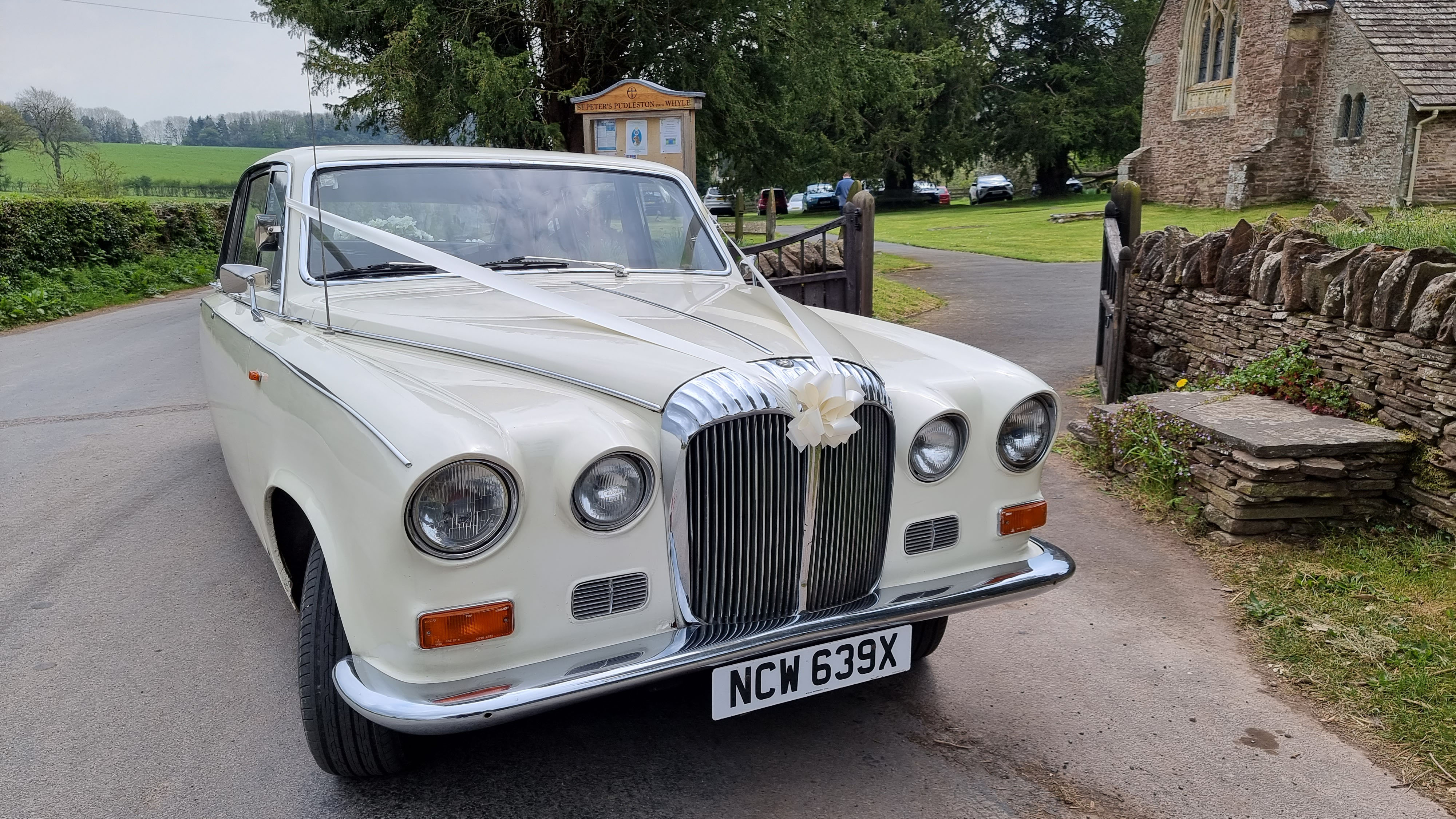 Front view of a cream classic Daimler limousine decorated with white wedding ribbons, parked outside a stone church.
