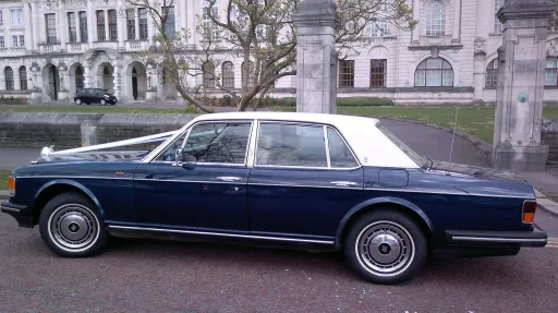Left side view of a blue Rolls-Royce Silver Spirit with a white vinyl roof, parked in front of historic buildings.