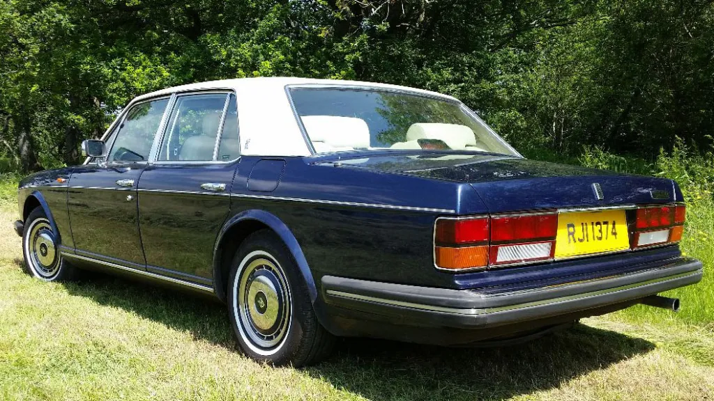 Rear view of a blue Rolls-Royce Silver Spirit with a white roof, showing chrome bumper and registration plate.