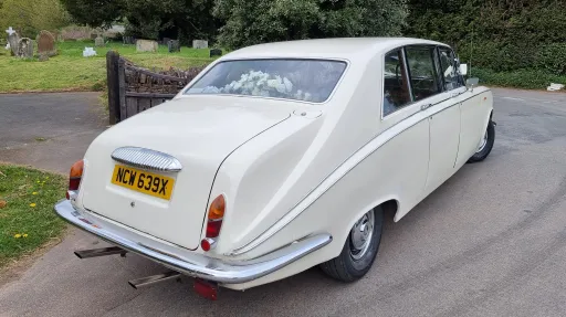 Rear view of a cream Daimler limousine parked on a roadside, showing chrome bumper and registration plate.