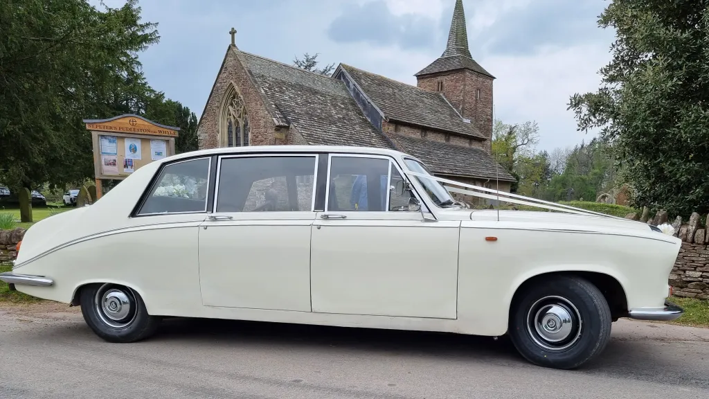 Right side view of a cream classic Daimler limousine decorated with white wedding ribbons, parked outside a historic stone building.