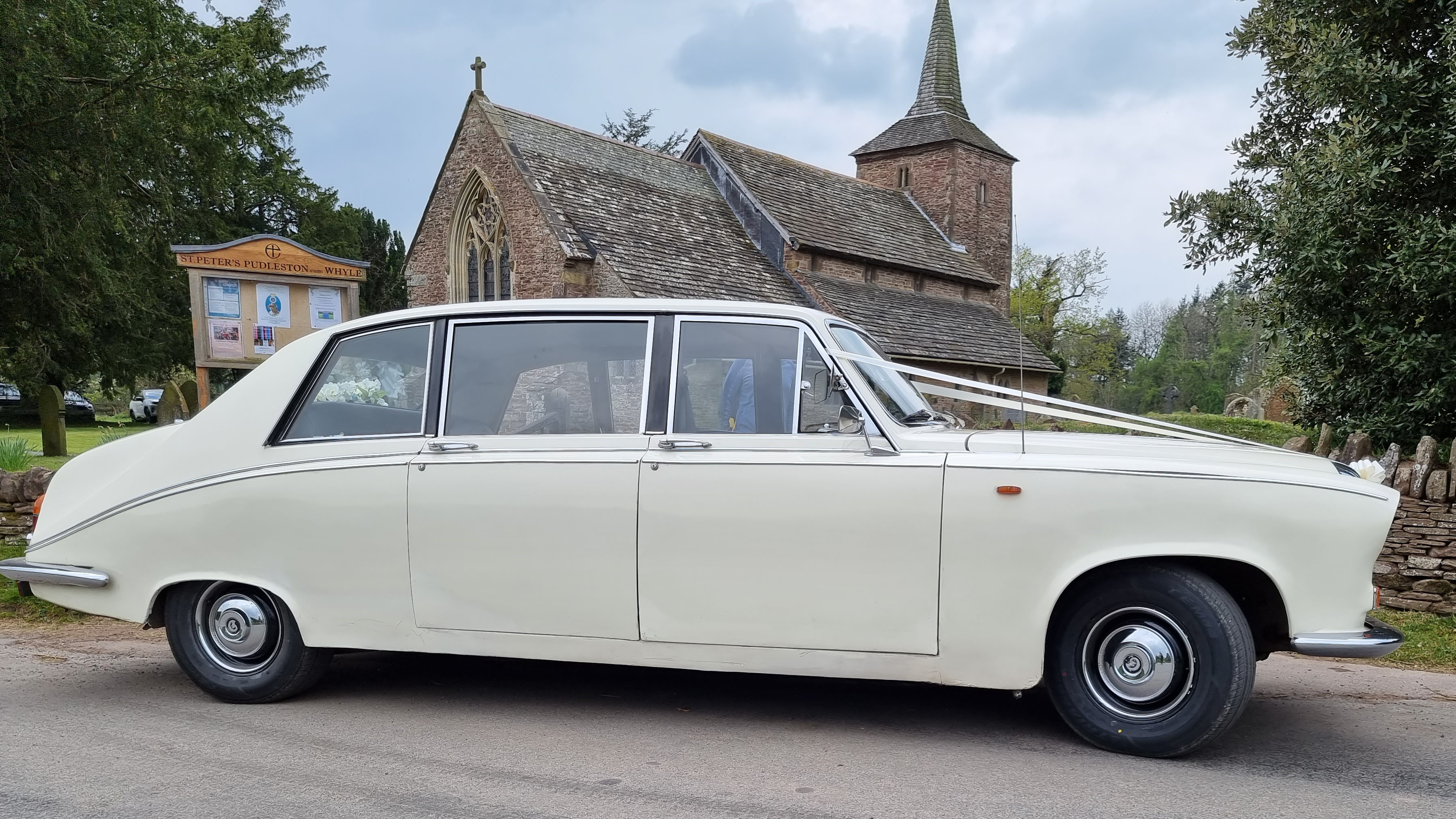 Right side view of a cream classic Daimler limousine decorated with white wedding ribbons, parked outside a historic stone building.
