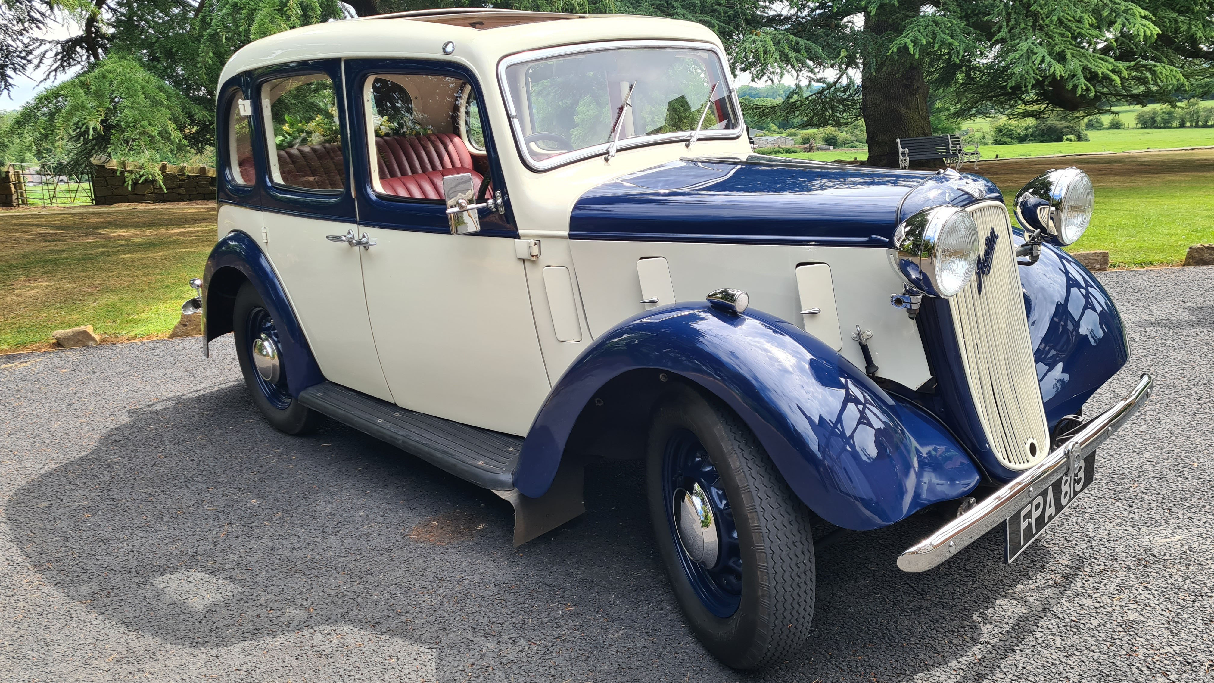 Front-angle view of a blue and ivory 1937 Austin 10 Cambridge on a driveway, showing its chrome grille, classic headlamps and vintage styling for wedding hire.