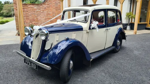 Side view of the blue and ivory 1937 Austin 10 Cambridge parked by a brick building, featuring its sunroof and two-tone bodywork, available for hire in Leicestershire.