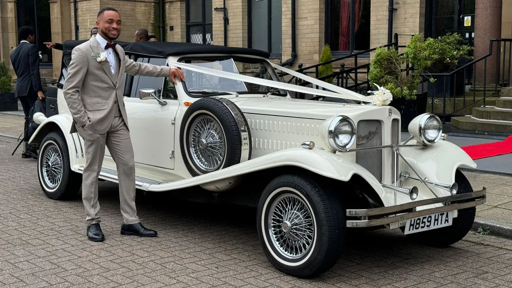 Groom posing next to an Old English White Beauford 4-door convertible wedding car at a West Yorkshire wedding venue