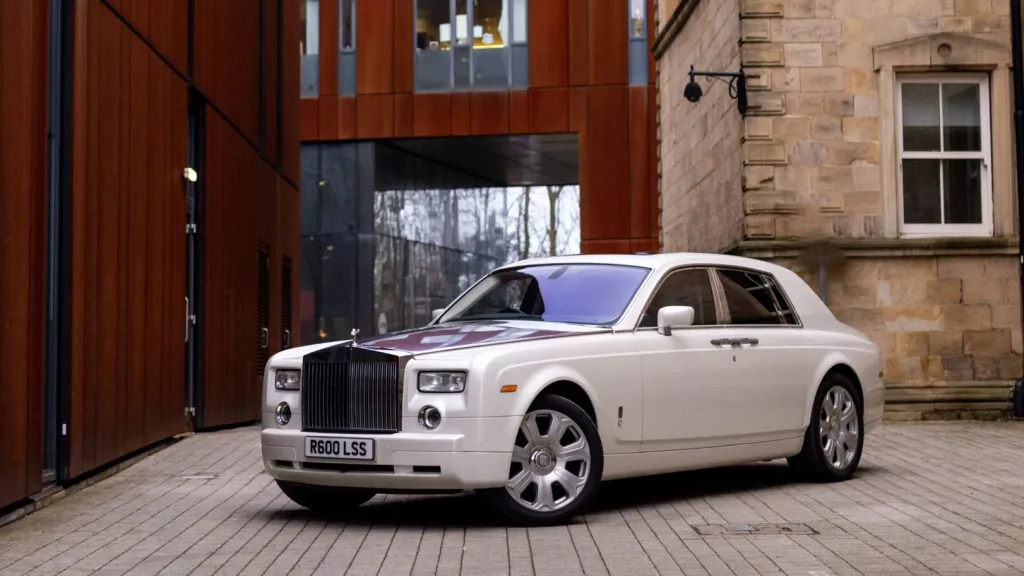 Front left side view of a white Rolls-Royce Phantom wedding car parked on a cobbled driveway outside a historic building