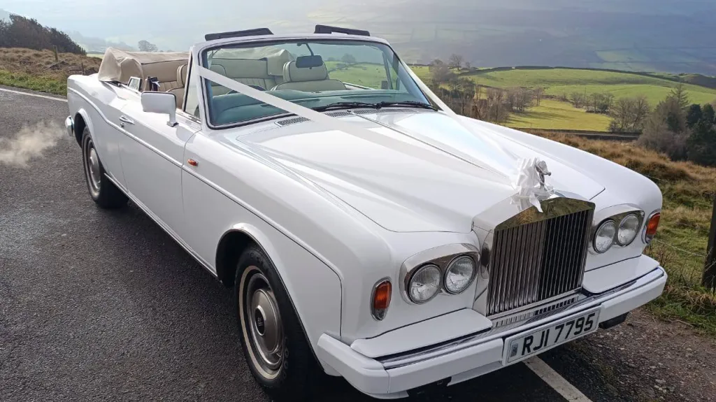 Front view of a white Rolls-Royce Corniche convertible wedding car with roof down, set against a countryside backdrop