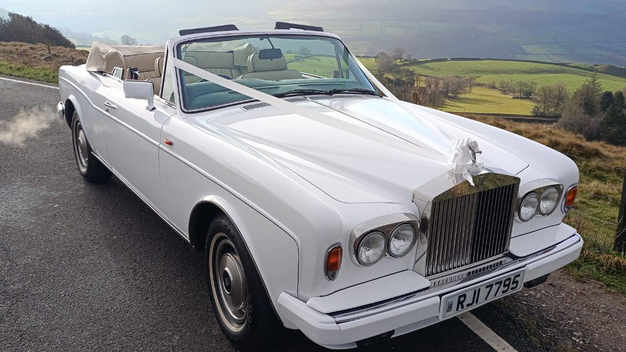 Front view of a white Rolls-Royce Corniche convertible wedding car with roof down, set against a countryside backdrop