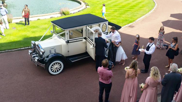 Aerial view of a cream 1930s style Imperial vintage wedding car surrounded by wedding guests outside the ceremony venue.